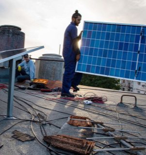 Professional solar installer carrying a blue solar panel during rooftop installation, showing cables and technical tools on a durable outdoor design project