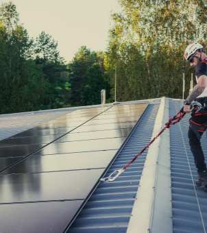 A professional technician with safety harness and helmet performing a solar system inspection and maintenance on a metal rooftop photovoltaic array.