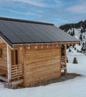 An off-grid wooden log cabin in a remote, snow-covered mountain area featuring a sleek, fully integrated BIPV solar roof tile system under a blue sky