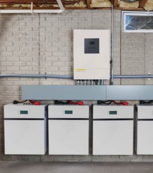 A real-life example of a residential solar energy storage system installed on a brick wall base. A row of four white wall-mounted home energy storage batteries are neatly connected in parallel at the bottom of the wall. Each battery has a thick red and black cable connected to its top, all neatly stored in gray metal cable trays. A beige photovoltaic inverter is installed directly above the batteries, surrounded by communication and power transmission pipes, showcasing the standardized installation of a modular home energy system.