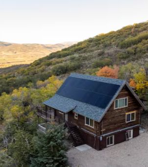 An aerial view of an off-grid log cabin in a remote mountainous area. Nestled beside a hillside covered in lush greenery and vibrant autumn foliage, a detached two-story American-style log cabin stands. The cabin features a dark wood structure, with one side of its gabled roof neatly covered with large black solar photovoltaic panels. In the distance, rolling desert-like hills and a clear sky vividly illustrate the practical application of off-grid energy systems in remote residences.