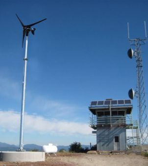 This is a real-world image of a green power supply system for a remote communication base station installed on a mountaintop. On the left, a towering white metal column stands, topped with a three-bladed black miniature wind turbine. In the center is a two-story base station equipment room with metal walls, its second-floor roof neatly arranged with multiple solar photovoltaic panels. On the right, a tall, grid-like metal communication tower stands, with two white microwave dish antennas mounted on its body. The background is a clear, bright blue sky with white clouds and distant mountain ranges, comprehensively showcasing the application of an off-grid wind-solar hybrid system at the base station.