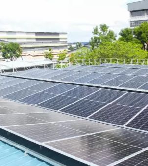 A real-life example of a rooftop solar photovoltaic (PV) power generation system in an industrial and commercial facility. Rows of large, dark black solar photovoltaic panels are neatly installed on the light blue corrugated steel roof of a building. In the mid-ground, green trees and metal railings are visible, while in the distance, other white and green industrial plants and commercial buildings, along with a cloudy sky, can be seen, showcasing the mature application of distributed rooftop PV in industrial parks.