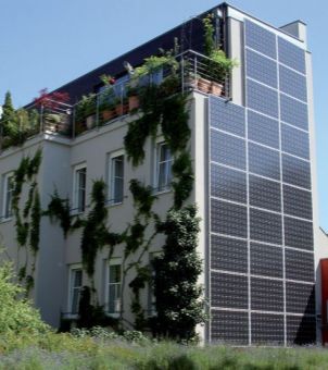 A real-life example of Building Integrated Photovoltaics (BIPV) in a modern green public building. The vertical facade of a multi-story modern building is covered with large, neatly arranged dark black solar photovoltaic panels. Green ivy and other plants climb the front facade, while the rooftop terrace is adorned with dense potted plants and metal railings. A green lawn in the foreground and a clear sky in the background vividly demonstrate the perfect integration of solar power generation and urban ecological architecture.