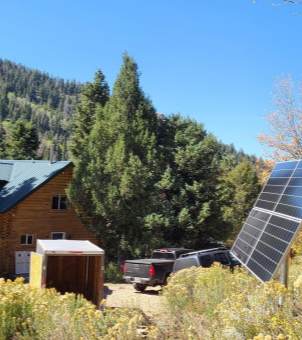 A remote, isolated log cabin stands against a backdrop of a clear sky and a densely forested hillside. Two pickup trucks are parked in front of the cabin, and in the foreground to the right stands a group of ground-mounted solar photovoltaic panels tilted towards the sun, surrounded by wild grass, vividly depicting a remote mountain dwelling's independent power supply via off-grid solar power.