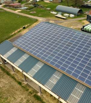 An aerial photograph taken from above. The main subject is the blue corrugated tile roof of a large building (such as an agricultural warehouse or residence), with its sun-facing slope neatly and densely covered with a large array of square solar photovoltaic panels. Surrounding the building is a vast expanse of green grass, dirt paths, and other agricultural facilities, clearly demonstrating the application scenario of using large-area rooftop photovoltaics for independent power generation and energy storage.