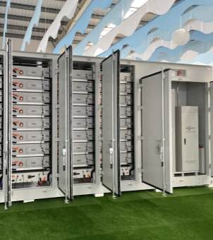 Inside an indoor showroom or factory, the floor is covered with green artificial turf, and above it is a unique blue and white wave-shaped decorative ceiling. The main focus of the image is a group of huge silver-gray industrial energy storage container cabinets, with multiple metal cabinet doors all open outwards. Inside the cabinets from the left to the middle, multiple layers of modular energy storage battery packs are neatly and densely stacked vertically, with complex black cables tightly connecting the batteries; the cabinet on the far right has a tall, gray-white electrical control cabinet (PCS converter system) installed independently, showcasing the internal structure of a large-scale industrial-grade energy storage device.