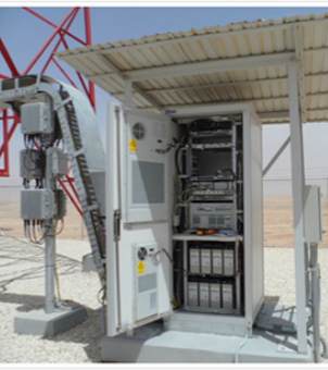 On the dry, hot desert or gravelly ground, a standardized communication base station cabinet equipped with a white metal rainproof and sunshade stands. The white door of the cabinet opens to the left. Inside, multiple layers of precision communication and power conversion electronic equipment are installed on the upper part, while two neatly arranged black backup battery packs are below. To the left of the cabinet, a white cable tray extends sloping down to the ground, with several gray junction boxes fixed on it. In the upper left background, a red communication tower structure is faintly visible.