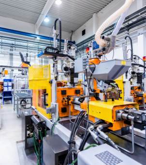 A spacious, bright, clean, and modern industrial manufacturing workshop. The scene shows neatly arranged complex automated machinery, painted in bright yellow and orange, topped with precision robotic arms, feeding hoppers, white exhaust hoses, and dense bundles of cable conduits. The workshop's ceiling is a high, white steel structure illuminated by numerous LED incandescent lights. In the background, other extended automated production lines and testing instruments can be seen, vividly showcasing an advanced industrial automation production scene.