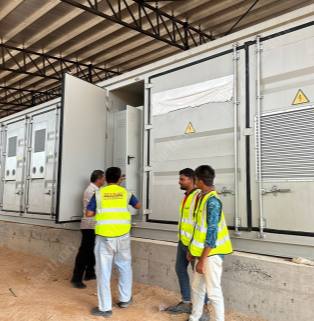 Real-world scene of on-site installation, commissioning, and operation and maintenance of containerized battery energy storage systems by technical personnel. Inside an industrial plant with a large steel-structured roof and earthen platform, a scene of on-site installation and operation of a containerized battery energy storage system is displayed. In the center of the image are neatly arranged large, standard white energy storage containers, securely placed on concrete bases. Yellow lightning bolt high-voltage danger triangle warning signs are printed on multiple inspection doors of the containers, and the right-side panel integrates large metal ventilation louvers. At this moment, one of the container's doors is fully open, revealing the gray-white internal control cabinet. Four male workers are gathered in front of the equipment: two are standing in front of the open door inspecting or discussing the equipment, while the other two young technicians, wearing bright yellow fluorescent safety vests, are talking on the right. The overall scene showcases the tense but orderly construction and commissioning atmosphere at an industrial-grade new energy storage project site.