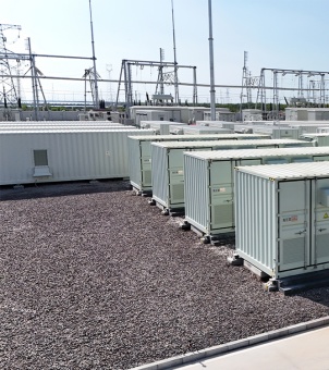 On a vast outdoor industrial site paved with gray gravel, rows of containerized battery energy storage systems are on display. In the foreground on the right is a unit consisting of five tall, closely spaced white energy storage cabinets, each topped with a dark gray square panel and fitted with a small black door lock assembly, all securely mounted on a white concrete base. Further along the gravel path extending to the left, more identical rows of white energy storage cabinets stand neatly in a line, one of which has its access door open, revealing glimpses of the complex gold and silver electrical components inside. In the midground on the left, a tall metal high-voltage transmission tower (grid tower) rises, above which a twilight sky, overcast with interspersed with vibrant golden rays of sunset, perfectly depicts the industrial park's energy infrastructure.