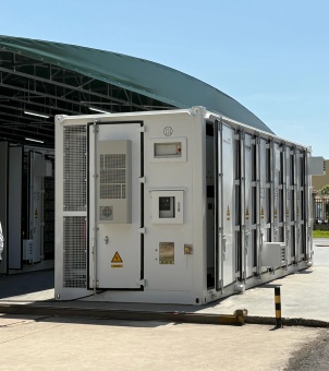 A close-up photograph of an outdoor commercial and industrial energy storage container under bright daylight. In the center of the image is a standard rectangular white energy storage container, independently installed on a concrete floor. Five double-leaf access doors, each equipped with sturdy metal locking bars and black sealing strips, are neatly arranged along its side. Small white temperature-controlled air conditioning units are partially mounted on the container. The front panel of the container is the core area for system control and power distribution, including an access door with metal mesh louvers (a bright yellow lightning bolt high-voltage danger triangle warning sign is affixed to the bottom of the door). The right side features an embedded glass display window, a separate square distribution box with switch valves and emergency stop components. To the left rear of the container is a semi-open factory building with a large green arched roof, through which another set of energy storage containers is faintly visible. To the right front stands a black and yellow safety bollard. The overall image presents a modern, industrial-grade power infrastructure system characterized by rigorous construction and explosion-proof safety.