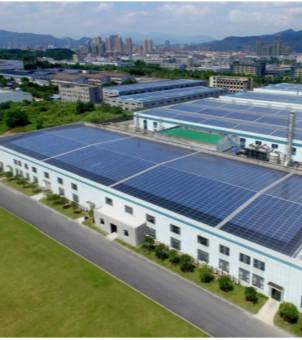 This is a wide-angle panoramic aerial photograph of a green, modern industrial park. The foreground and middle ground feature several massive, elongated, modern white industrial buildings with multiple windows. The flat roofs of these buildings are entirely covered with large arrays of deep blue solar photovoltaic panels, shimmering in the sunlight. A smooth black asphalt road runs alongside the buildings, lined with neatly planted green spherical trees, connecting to a spacious green lawn demonstration area. In the background, the view extends beyond the low-rise factory buildings to a dense cluster of city skyscrapers on the distant horizon, and further still, rolling green mountains. The sky is pale blue with wispy white clouds. The photograph's expansive view and distinct color layers vividly showcase the grand ecological scene of the industrial park's large-scale application of photovoltaic and energy storage clean energy.