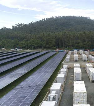 This is a panoramic outdoor photograph taken from above, showcasing a large-scale, centralized ground-mounted photovoltaic (PV) power generation and containerized energy storage power station. On the left is a vast plain, with rows of long, dark blue ground-mounted PV panels arranged neatly and densely, stretching parallel into the distance. On the right, the ground has been hardened and paved with gravel, neatly divided into several longitudinal channels. Two rows of more than ten large, light-colored containerized lithium-ion battery storage units and converter compartments are parked in a standardized manner, each container equipped with a lifting structure and heat dissipation module on top. In the background of the entire PV-storage base is a massive, towering hill covered with lush, dark green trees, above which is a pale blue sky dotted with large areas of layered white clouds. The entire image demonstrates a modern, mega-scale PV-storage complementary project that integrates new energy infrastructure with the natural environment.