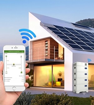 This is a high-tech promotional image showcasing a smartphone app for remote monitoring of a residential green solar energy storage system. In the foreground on the left, a hand holds a white smartphone, its screen clearly displaying the green and white energy management app interface, including a complex power flow topology diagram, battery percentage, and a data list below. Above the phone, three progressively larger blue Wi-Fi signal ripple icons are blurred and overlaid. The background is a modern, white, irregularly shaped detached villa with a wide, sloping roof covered in deep blue solar photovoltaic panels. The villa's ground floor features large, floor-to-ceiling windows, emitting warm yellow light. On the outdoor ground floor to the right of the villa, a white, vertical, modular energy storage unit is placed, also topped with a small blue Wi-Fi signal icon. The entire image cleverly combines the software interface with the real-world architecture, vividly conveying the convenience of smart home energy management.