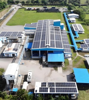 This is a panoramic aerial photograph taken from a high vantage point, showcasing an industrial park featuring a large-scale commercial and industrial rooftop distributed photovoltaic and energy storage integrated project. At the center of the image is a towering, modern manufacturing plant with a dark blue steel-framed flat roof. Thousands of densely packed, dark black solar photovoltaic panels are neatly arranged on its massive rectangular roof, with passageways for lighting and maintenance in the center. Several connecting corridors extend from the left and right sides of the main plant, interspersed with smaller white auxiliary buildings, office bungalows, and carports, all with their blue roofs also extensively covered with photovoltaic panels. The ground in the plant area is mostly paved with gray-white cement, with several white trucks and logistics vehicles parked there. The complex is surrounded by large, neatly trimmed, bright green lawns and dense trees, allowing bright natural light to penetrate. The overall layout is grand, clean, and tidy, clearly demonstrating the modern scale of this 1.7MW-level commercial and industrial photovoltaic and energy storage integrated demonstration project.