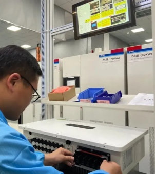 This is a real-life photo of an industrial laboratory showcasing the rigorous testing and quality control process of hybrid inverters by technicians. In the foreground on the left, a young male technical expert wearing a blue anti-static cleanroom suit and glasses is focused on his work. His hands are adjusting the wiring and control interfaces on the lower front of a white rectangular hybrid inverter placed flat on a workbench. Two rows of round black terminals are neatly arranged on the left side of the inverter. On a multi-tiered display shelf above the workbench, a kraft paper box containing red components and two plastic material boxes, one blue and one dark blue, each containing small components, are neatly arranged. In the background, large white power testing cabinets are neatly arranged, with prominent red and blue labels on top, clearly indicating the test source information, such as 'AC source (75KVA)' and 'DC source (80KW)', in black Chinese and English font. A tilted black LCD monitoring screen hangs directly above the entire testing station, displaying complex electrical system operation diagrams and yellow warning boxes. The entire image exudes the atmosphere of modern power electronics R&D and rigorous product testing within an industrial technological environment.