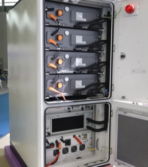 A close-up photograph of the actual structure of an integrated off-grid energy storage system combining photovoltaic, energy storage, and diesel power, taken at an industrial exhibition. The main subject of the image is a vertical, white, integrated steel energy storage cabinet. Both upper and lower cabinet doors on the right side are fully open, clearly revealing the intricate hardware layout of the upper and lower compartments. In the upper cargo compartment, four identical dark gray cuboid battery modules are stacked vertically and compactly. Each module has a bright orange safety high-voltage electrical connector on its left front, leading to a thick black cable that converges vertically downwards. The lower cargo compartment houses an independent electrical control unit, containing a gray converter control main control chassis with a large black LCD display panel. Below this is a complex array of switches, red and green terminals, and four miniature black cooling fans arranged side-by-side. A prominent circular red emergency stop button is mounted on the top inside the open white cabinet door on the right, while a large gray honeycomb metal dustproof and heat dissipation mesh is embedded in the lower cabinet door. The server rack is placed on a purple exhibition stand, with the blue carpet and modern architectural structure of the exhibition hall faintly visible in the left background. The entire image exudes a sense of technology and exquisite craftsmanship from high-performance, multi-energy complementary industrial energy storage equipment.