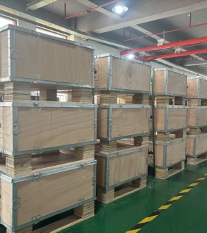 This is a real-life photo showcasing a large quantity of energy storage equipment, neatly stacked in heavy wooden crates, ready for export in a modern industrial warehouse. The image shows several towering packaging matrices arranged vertically from near to far, each matrix consisting of three large, light yellow, multi-layered heavy wooden crates stacked vertically. These crates are meticulously crafted, with thick, silver-plated galvanized steel strips and fasteners reinforcing their edges and sides, demonstrating a high standard of impact protection for export packaging. The bottoms of the crates are integrated with thick, multi-layered solid wood pallet legs, facilitating forklift handling. The entire matrix of crates is neatly arranged on a clean, dark green epoxy floor, marked with prominent yellow and black diagonal safety warning lines. Above, the high ceiling is fitted with long rows of gray cable trays, red fire hydrants, and bright industrial lighting fixtures. The entire warehouse environment is orderly and exudes the industrial atmosphere of modern mass manufacturing and export logistics.