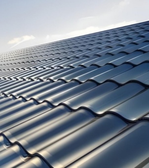 This is a photograph taken from a slightly downward angle, showcasing a modern residential building-integrated photovoltaic (BIPV) solar roof tile installation. The main subject is a large expanse of deep blue solar photovoltaic roof tiles extending to the upper right, featuring a traditional and elegant wave-like design reminiscent of Chinese glazed tiles. Under the gentle natural sunlight, the smooth, mirror-like deep blue glass surface of the tiles shimmers with a subtle water-like sheen and a delicate metallic luster, demonstrating high-standard rainproofing, waterproofing, and integrated photovoltaic encapsulation technology. A faint blue sky and wisps of white clouds peek through the edge of the roof, creating a clean and serene scene that clearly presents the visual effect of green building materials deeply integrating cutting-edge photovoltaic technology with residential aesthetics.