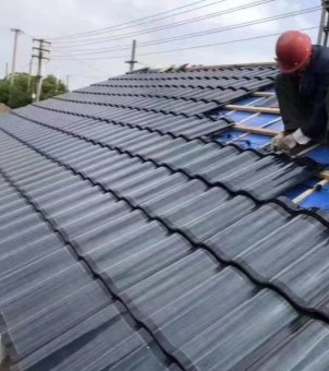 This is a real-life photo showcasing the on-site construction and installation of modern Building Integrated Photovoltaics (BIPV) residential solar roof tiles. The main subject is a sloping residential roof under construction, presented in a perspective composition extending from the lower left to the upper right: the left side of the roof is largely covered with rows of dark black, smooth-surfaced solar photovoltaic tiles with elegant wavy lines; the upper right shows the unfinished construction area, clearly revealing the bright blue waterproof base layer, the crisscrossing light yellow wooden battens, and the vertically arranged electrical wiring. In the upper right corner, a construction worker in dark overalls, wearing a red hard hat and gloves, is crouching on a wooden scaffold, focused on securing the photovoltaic tiles and connecting the cables. Above the roof, the dense network of overhead power lines and the overcast sky are faintly visible. The entire scene is realistic, full of industrial construction texture, vividly presenting the engineering details of solar tile installation from the basic framework to the finished product.