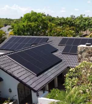 This is a bird's-eye view photograph showcasing the ecological landscape of a modern detached house with a solar photovoltaic system installed on its roof. The main subject is a white-walled, gray traditional wave-patterned tiled roof with a staggered roof structure. On the three sun-facing slopes, three neatly and compactly installed arrays of all-black rectangular monocrystalline silicon solar panels (one in the upper left, one in the foreground, and one on the right), the black panels harmonizing with the gray roof tiles. A retro-style stone chimney built from natural stones stands beside the right eaves, and a black retro wrought iron gate and white pillars are faintly visible at the ground floor entrance. The house is surrounded by lush, verdant tropical and subtropical trees and shrubs, with a backdrop of a vast green forest on the horizon and a bright sky dotted with wisps of white clouds. The entire scene, bathed in sunlight and lush greenery, vividly presents the integration of a home's distributed solar power with the natural environment.