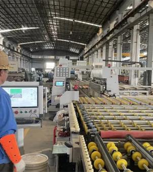 This photograph showcases a modern industrial factory's automated production line for the mass production of solar roof tiles. The image employs a deep perspective composition extending from left to right: in the foreground on the left, a technician in blue overalls, a bright orange cut-resistant arm guard on his left arm, and a yellow safety helmet stands sideways, facing a modern control panel equipped with a digital LCD screen and control switches. The screen displays green production status and monitoring feeds. Extending to the right rear from the foreground is a massive and densely packed automated roller conveyor line, with countless golden round conveyor rollers neatly arranged on metal tracks. In the midground stand several large, white, fully automated precision machine tools and CNC equipment, each with a small control panel and blue indicator lights. The background is an extremely tall and spacious steel-structured industrial plant with a black, arched, perforated metal roof covered with longitudinal beams and massive concrete supporting columns. Bright natural light streams in through the factory gate in the distance. The entire scene was well-organized and large-scale, fully demonstrating the intelligent automated processing and high-efficiency assembly line production of new energy building materials.