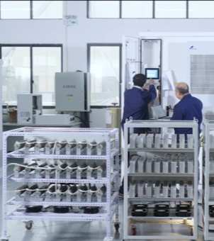 This is a real-life industrial scene photo showing engineers conducting software and hardware collaborative system debugging inside a new energy storage and converter R&D laboratory. In the foreground, two multi-layer metal test racks equipped with casters are placed side by side: on the left is a white four-layer angle steel rack, each layer of which is arranged horizontally in a very tight and orderly manner with two rows of dark-colored columnar electrical components with metal terminals, and the bottom layer of the rack is equipped with a row of black circular cooling fans; on the right is a similar gray five-layer rack, each layer of which is densely lined with dozens of silver-white rectangular modular electronic components or metal capacitors, and the bottom layer is also equipped with six small black fans arranged side by side. In the mid-range shot behind the shelving, two male technical experts in dark blue long-sleeved uniforms are facing away from the camera, operating a large white high-voltage converter inverter cabinet. The thinner engineer on the left raises his right hand and clicks on a rectangular digital LCD touchscreen display emitting a bright blue light inside the cabinet door; the heavier engineer on the right is slightly turned to the side, holding an open, silver-gray, thin laptop in both hands, seemingly monitoring and recording debugging data simultaneously. The laboratory backdrop is a tall, bright white factory wall, with several neatly arranged, high-rise, black-framed glass windows for natural light. At the far end, a gray electrical device with a yellow high-voltage warning sign sits on a table. The entire scene is orderly and exudes a rigorous atmosphere of modern green energy research and lean engineering technology.