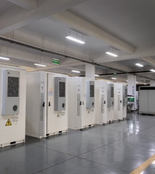 This photo, taken from a slightly angled front, showcases a modern industrial factory where a large number of integrated energy storage cabinets are neatly arranged and ready for shipment. The composition employs a strong perspective extending from left to right: on a smooth, gray epoxy floor, a row of dozens of pure white rectangular industrial and commercial distributed energy storage cabinets stands in perfect order. Each white cabinet has a long, light gray dedicated industrial air conditioning unit vertically mounted on its front door, with a circular metal exhaust fan shroud on the lower half. A prominent yellow triangular high-voltage hazard warning label with a lightning bolt symbol is affixed to the lower part of the cabinet door. The sides of the cabinets feature long, louvered ventilation grilles and black fastening handles. Above, a tall, spacious white concrete load-bearing ceiling is illuminated by multiple long, parallel, high-brightness LED industrial lights, making the entire workshop bright and airy. Green safety signs hang below the ceiling. The entire scene was well-organized and large-scale, fully demonstrating the mature capabilities of new energy hardware manufacturing in large-scale mass production, spot inventory, and commercial supply chain delivery.