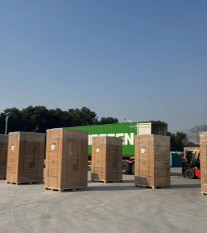 This photo, taken on a clear day, shows a large-scale global logistics shipment of new energy industrial and commercial energy storage cabinets or large electrical equipment being loaded into containers. In the foreground, on a concrete plaza, at least five sets of tall, rectangular cabinets are neatly arranged, each wrapped in a sturdy brown industrial cardboard box with a fumigation-free wooden pallet at the bottom for easy forklift handling. The entire shipment is covered with multiple layers of dense, reflective, transparent waterproof plastic wrap. The sides of the boxes are faintly printed with compliant logistics markings, handling labels, and yellow high-voltage warning triangles. In the mid-ground, a long container semi-trailer truck is parked horizontally, carrying a striking bright green Evergreen Marine international standard shipping container with a large white 