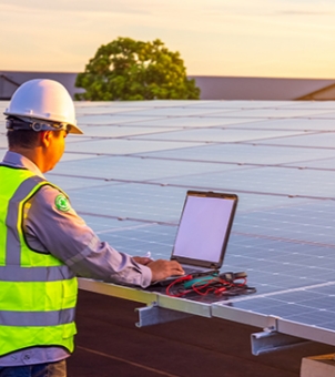 This photograph, taken in the warm light of a sunset, depicts a real-life workplace scene of photovoltaic installers and EPC company engineers conducting on-site grid connection commissioning and operation and maintenance of a rooftop solar power station. In the foreground on the left, a professional technician wearing a gray long-sleeved work uniform, a bright fluorescent yellow safety vest with gray reflective strips, and a white standard safety helmet stands intently to the side. His hands are resting on the keyboard of a black portable laptop placed on the edge of the photovoltaic modules; the computer screen is slightly lit, and below the keyboard are scattered professional electrical testing instruments (such as multimeters or string testers) connected with red and black test leads. In the middle ground, a large, modern distributed solar photovoltaic panel array stretches out to the right rear. Blue monocrystalline silicon photovoltaic modules are neatly fixed on silver-gray aluminum alloy or steel structure support tracks at the bottom, their surfaces reflecting the gentle golden afterglow of the setting sun. In the background, a lush green tree stands out, and the rooftops of other buildings are faintly visible in the distance. The entire sky was bathed in soft pale orange and light yellow hues by the setting sun, creating a vibrant and clear picture that vividly showcased the rigorous and professional digital engineering delivery and on-site technical support during the later stages of the new energy power plant construction.