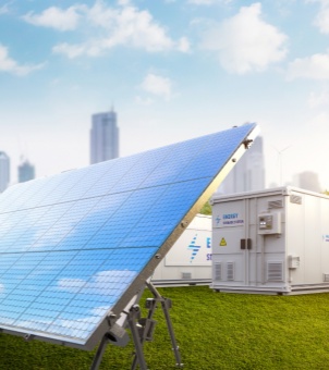 This image, rendered in high-definition 3D under a bright, clear blue sky, showcases a technological concept of an integrated solar and storage solution. In the foreground, a large array of monocrystalline silicon solar panels is mounted diagonally on the left, their deep blue, grid-like reflective surfaces clearly reflecting the sky and clouds above. In the middle ground, a lush, green lawn stretches out. To the right, two pure white, standard containerized integrated energy storage (ESS) units are arranged side-by-side. Each unit features double security doors and ventilation grilles, with the words 'ENERGY STORAGE SYSTEM' printed on the front top with a blue lightning bolt logo. A yellow triangular high-voltage lightning safety label is affixed to the side of each unit. The background, bathed in bright, gentle sunlight, presents the hazy silhouette of a modern metropolis skyline in the distance, while a tall, white wind turbine is faintly visible in the background on the right. The image is vibrant in color and has a layered composition, vividly showcasing the environmentally friendly technological landscape of modern cities empowered by green distributed solar power generation and container energy storage systems for low-carbon development.
