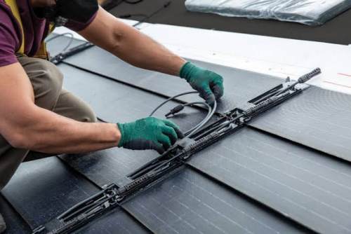 On-site Installation and Wiring of Roof Photovoltaic Tiles A close-up, realistic photograph shows an installer wearing green non-slip work gloves and brown work pants kneeling on a black roof, carefully arranging and connecting gray cables and waterproof connectors between the photovoltaic roof tiles. The black solar photovoltaic tiles are neatly arranged on the roof, their slotted structure and internal wiring channels clearly visible.